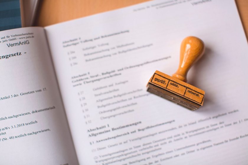 A wooden verification stamp resting on an open book of statutes, its face reading geprüft, sachlich, rechnerisch